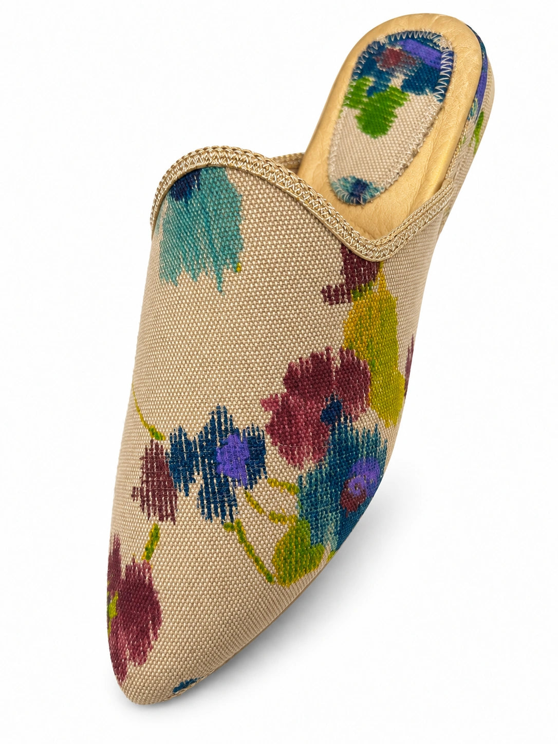 Beige woven babouche with blue, burgundy and purple flowers, green leaves and pale braided edging on a white background.