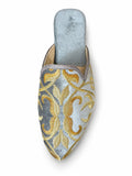 Pale blue-grey babouche with gold scroll motifs and a pointed toe on a white background.